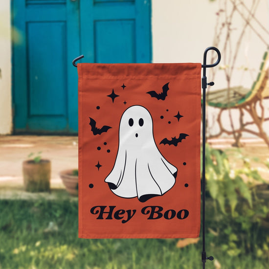 Halloween garden flag featuring a cute white ghost with black bats and stars on an orange background, with the phrase 'Hey Boo' in black, perfect for festive outdoor decor.