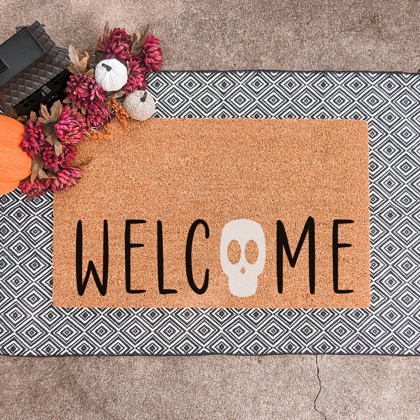 Halloween doormat with the word 'Welcome' featuring a skull as the letter 'O,' displayed on a geometric black and white layering rug with decorative pumpkins and flowers.