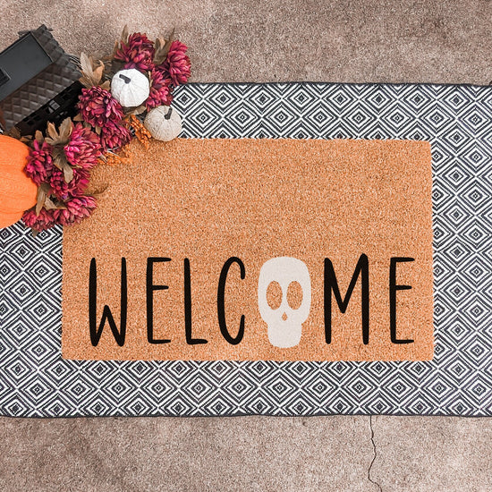 Halloween doormat with the word 'Welcome' featuring a skull as the letter 'O,' displayed on a geometric black and white layering rug with decorative pumpkins and flowers.