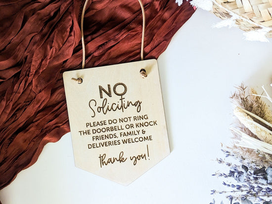 No Soliciting sign with engraved text advising not to ring the doorbell or knock, displayed on a red fabric backdrop with dried flowers, perfect for keeping a peaceful entryway.