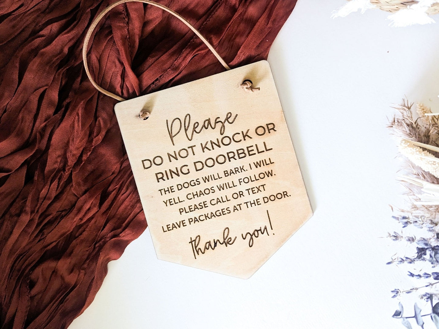 Please Do Not Knock or Ring Doorbell sign with humorous engraved text about barking dogs and chaos, displayed on a red fabric backdrop with dried flowers, perfect for pet-friendly households.