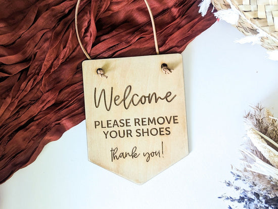 Welcome sign with engraved text reading "Please Remove Your Shoes, Thank You!" displayed against a red fabric backdrop, ideal for entryway or home decor.