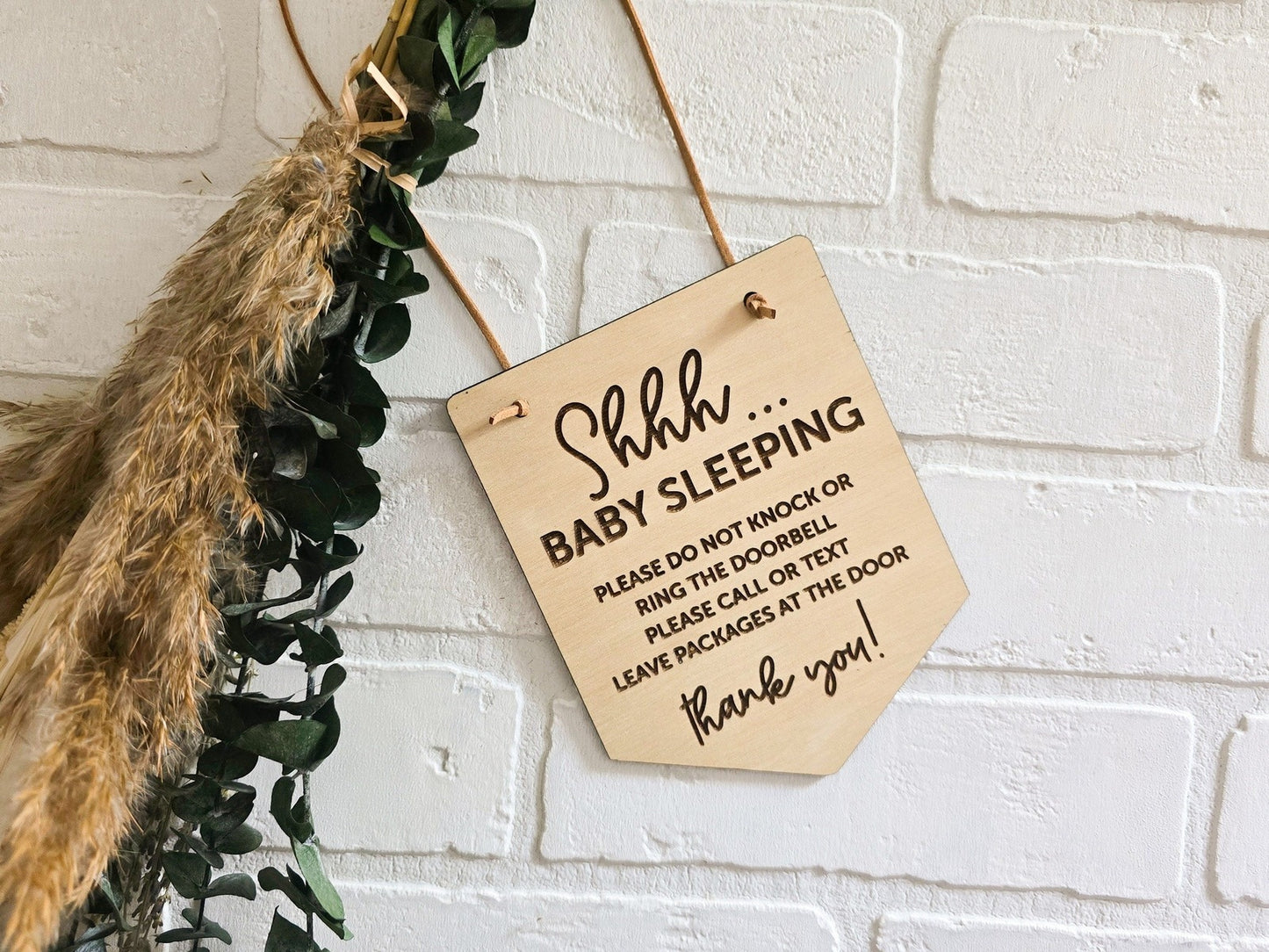 Shhh... Baby Sleeping sign with engraved text advising not to knock or ring the doorbell, displayed against a white brick background with decorative greenery, perfect for quieting the entryway of a home with a sleeping baby.