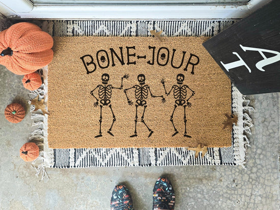 Bone-Jour doormat featuring three dancing skeletons, displayed on a fringed layering rug, surrounded by decorative pumpkins, perfect for Halloween porch decor.