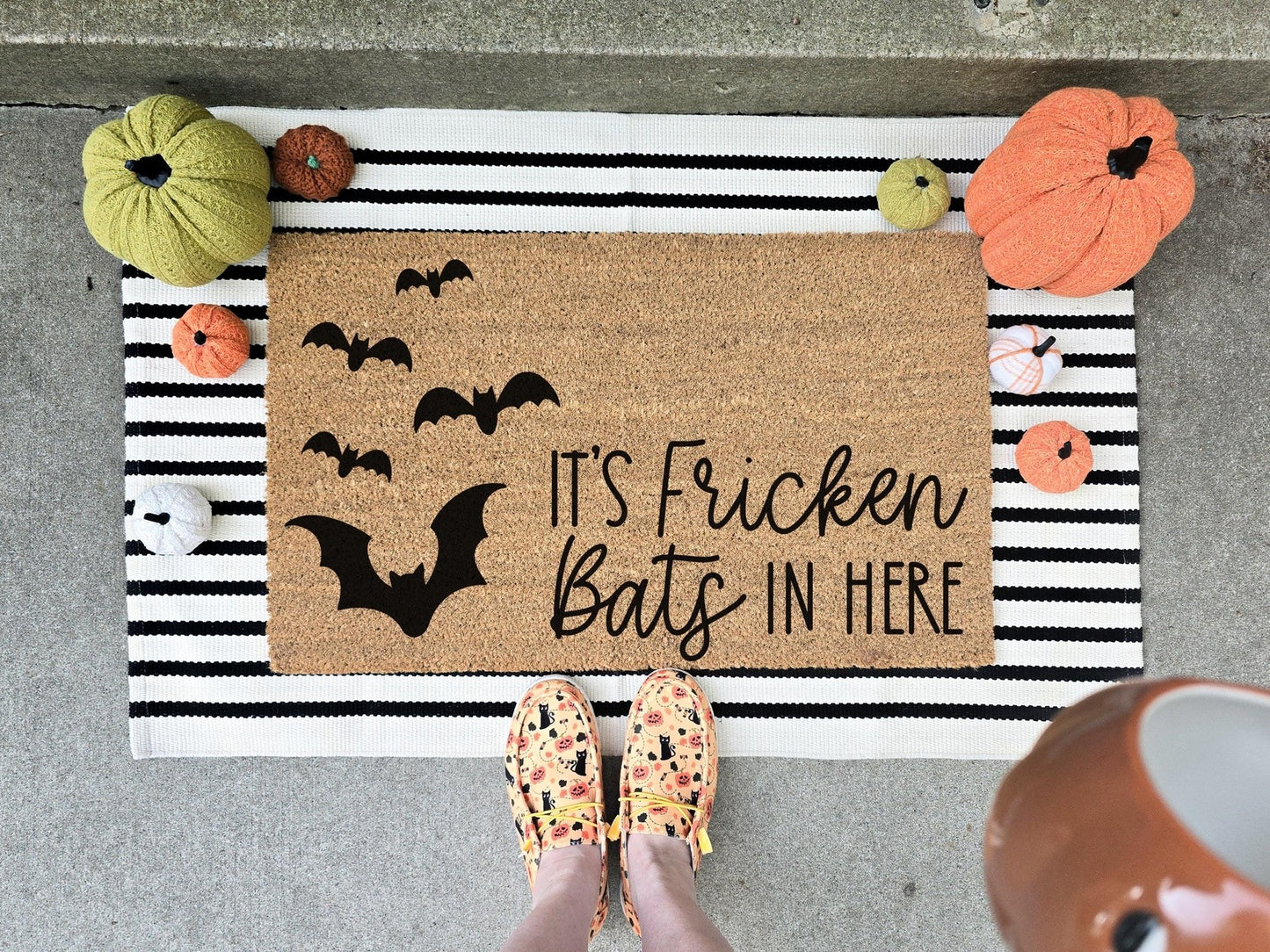 It's Fricken Bats In Here doormat featuring a cluster of black bat designs, displayed on a striped layering rug with decorative pumpkins, perfect for Halloween porch decor.