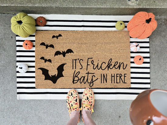 It's Fricken Bats In Here doormat featuring a cluster of black bat designs, displayed on a striped layering rug with decorative pumpkins, perfect for Halloween porch decor.