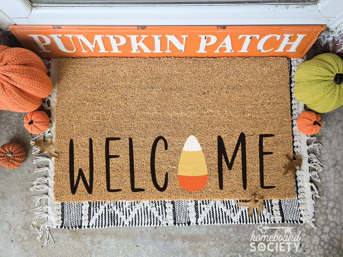 Fall-themed doormat with the word 'Welcome' featuring a candy corn as the letter 'O,' displayed on a fringed layering rug, surrounded by decorative pumpkins and a 'Pumpkin Patch' sign, ideal for autumn porch decor.