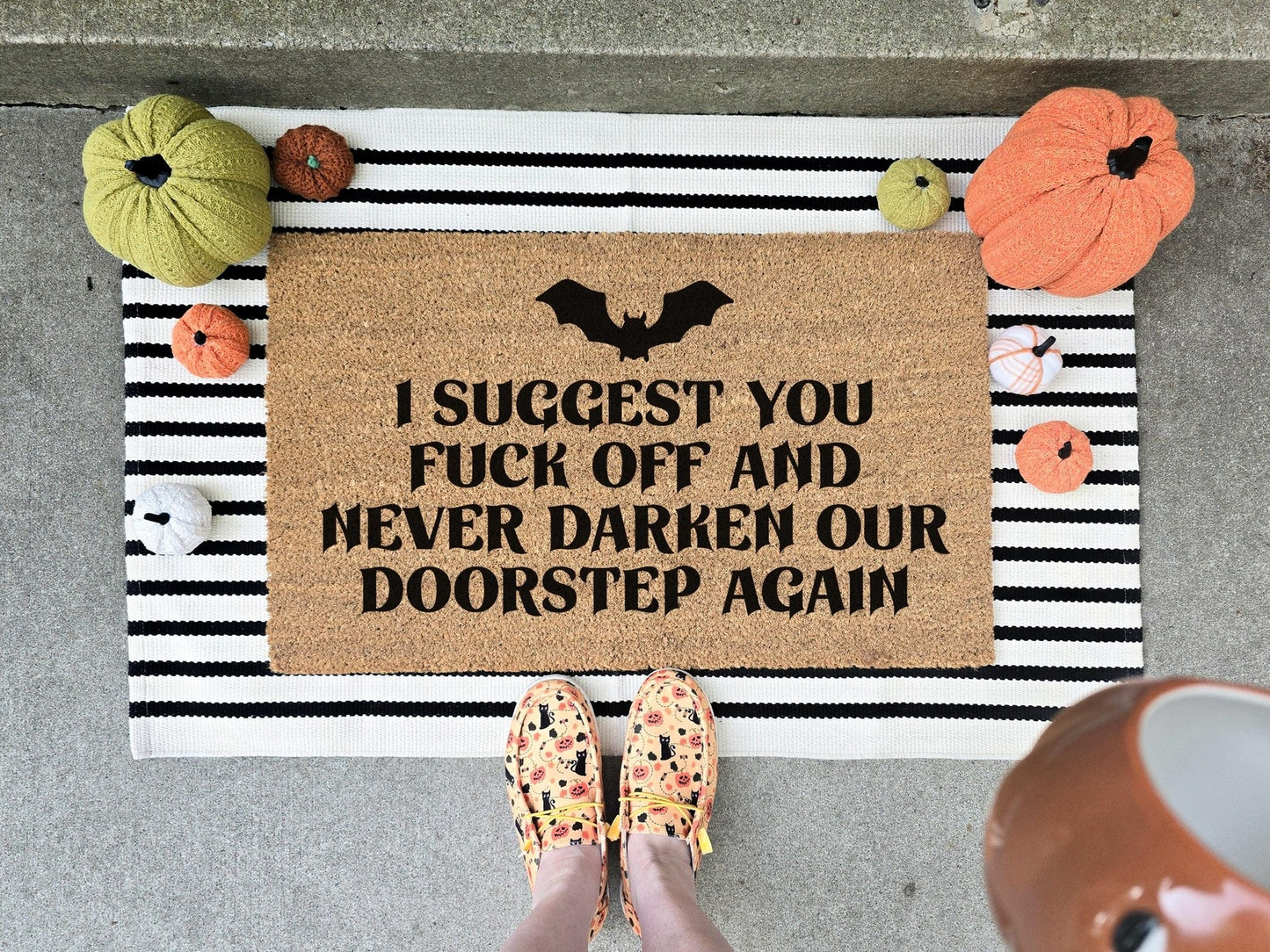 Humorous Halloween doormat with bold text that reads 'I suggest you fuck off and never darken our doorstep again' featuring a black bat design, displayed on a striped layering rug with decorative pumpkins.