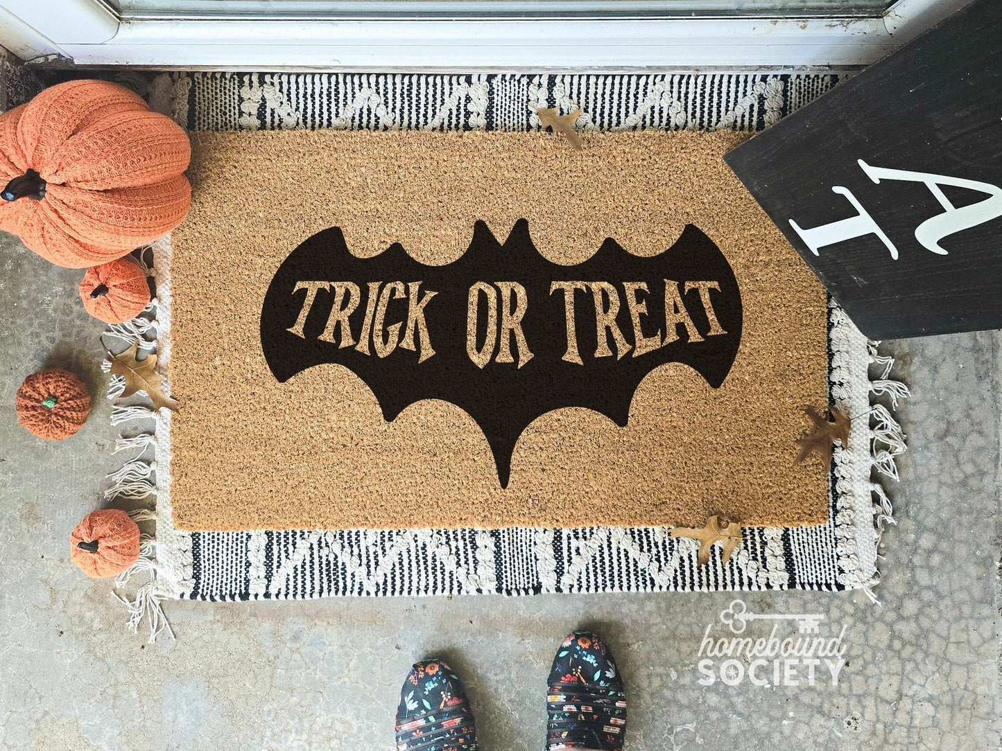 Halloween doormat with a black bat design featuring the text 'Trick or Treat,' displayed on a fringed layering rug, surrounded by decorative pumpkins, ideal for spooky outdoor decor.