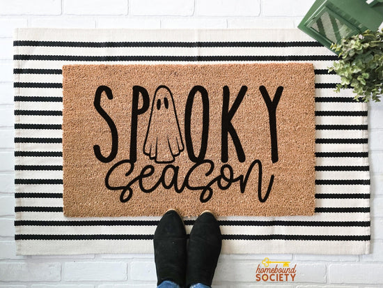 Hand-painted 'Spooky Season' doormat featuring a ghost design, displayed on a black and white striped layering rug, perfect for Halloween-themed front porch decor.